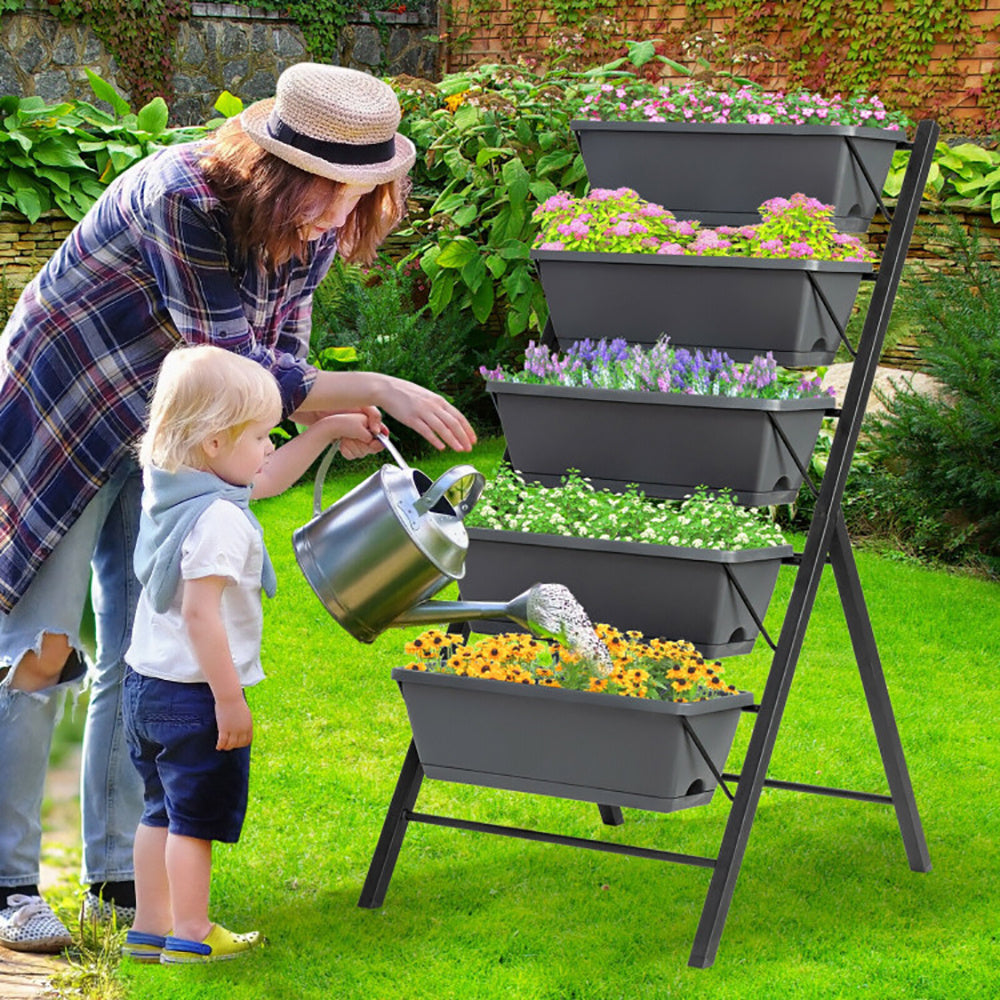 Coocosh tiered planter stand with a woman and child watering flowers. The dark gray planter is made of plastic, featuring five levels of planters filled with yellow and pink flowers, ideal for small garden spaces. Outdoors perspective. 