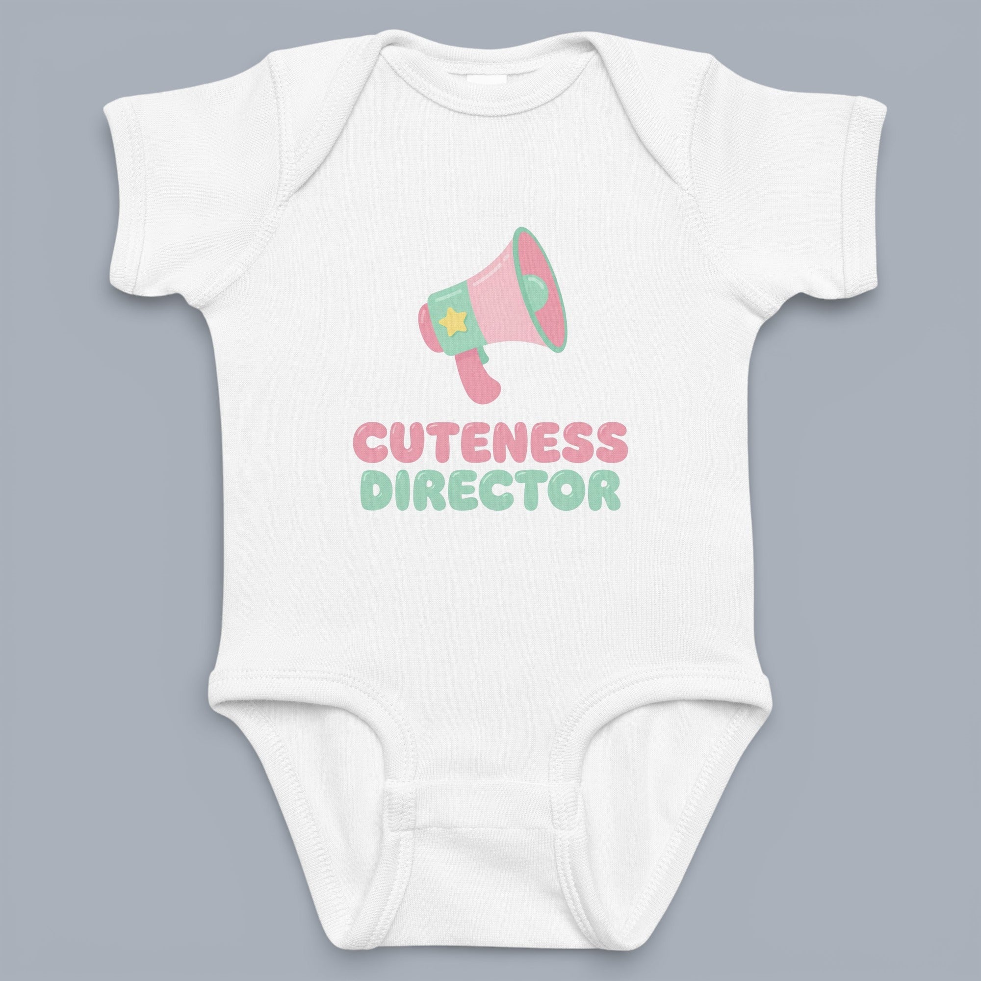 Coocosh baby onesie featuring a cute megaphone graphic, with the text "Cuteness Director" in pink and green. Soft, breathable fabric, short sleeves, white color. Adorable for your little one's wardrobe. A fun and playful design. Perfect for everyday wear 