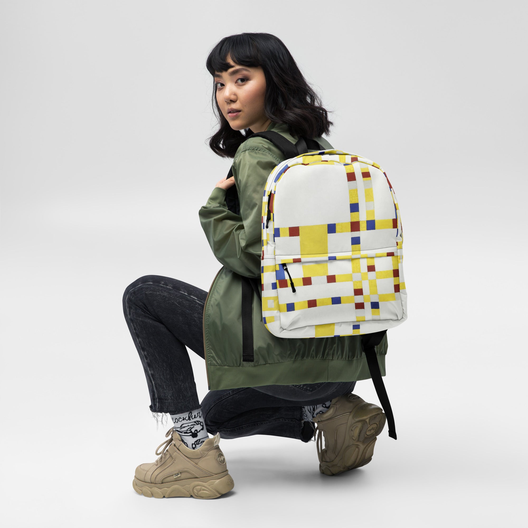 Coocosh's Broadway Boogie Woogie geometric abstract backpack, worn elegantly in a crouching pose, showcases vibrant yellow and white patterns. Designed for daily adventures, its ergonomic straps and spacious laptop compartment ensure style meets functionality.